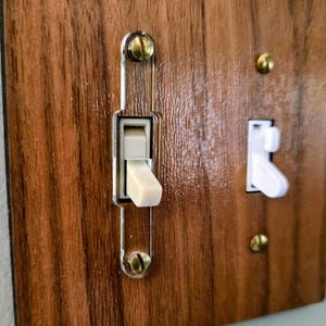 May include: A rectangular light switch wall plate with a faux wood grain finish. The plate features two toggle switches, one beige and one white, secured with brass-colored screws. A clear acrylic frame surrounds the beige toggle switch.