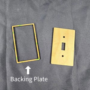 Könnte beinhalten: Eine Holz-Lichtschalterabdeckung und eine Rückplatte. Die Rückplatte ist rechteckig mit einem grünen Kleberand. Die Schalterabdeckung ist rechteckig mit einer einzigen Schalteröffnung und zwei Schraubenlöchern. Der Text "Backing Plate" befindet sich darunter.