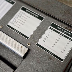 Op de afbeelding: Een grijs getextureerd metalen oppervlak met een zilveren handvat en twee informatieve stickers. E&eacute;n sticker is getiteld Wood Glue Guide, de andere is een Pilot Hole Sizes tabel voor houtschroeven, met maten van #4 tot #14 in inches.