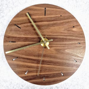 May include: A round wooden wall clock with a dark brown finish. The clock features gold-colored hands and markers. The clock's face has a natural wood grain pattern. The clock measures approximately 12 inches in diameter.