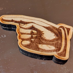 May include: A wooden cutout of a hand pointing to the right. The hand is intricately detailed with etched lines and shading, giving it a realistic appearance. The edges are dark, creating a contrast with the lighter wood grain.