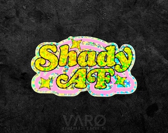 May include: A die-cut sticker featuring the text Shady AF in bold yellow letters with a holographic finish. The text is outlined in pink and accented with small yellow star shapes against a shimmering, multi-colored holographic background.