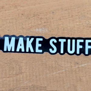 May include: Black and white sticker with the text "MAKE STUFF" in a bold, sans-serif font. The sticker has a black background with a white border and text. The words are in all capital letters. The sticker also includes the text "THE NEW JANET WORKSHOP".