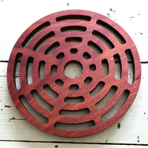 May include: Circular wooden trivet with a reddish-brown finish. The trivet features a series of concentric rings with cut-out sections, creating a decorative and functional design. The trivet is designed to protect surfaces from hot items.