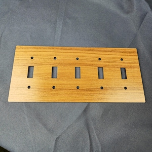 May include: A rectangular light brown wooden switch plate with a wood grain pattern. The plate has four rectangular openings for switches and eight small circular holes for screws. The plate is designed to cover multiple switches.