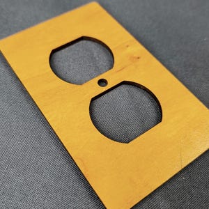May include: A rectangular light brown wooden electrical outlet wall plate featuring two rounded openings for a duplex receptacle and a small centered hole for a mounting screw, showcasing a smooth finish and natural wood grain patterns.