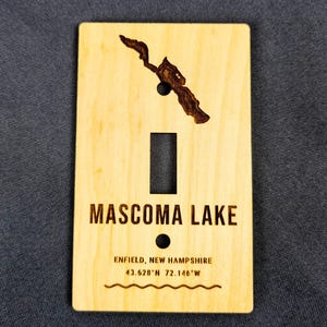 May include: A rectangular light-colored wooden light switch cover plate featuring a dark laser-engraved map of Mascoma Lake at the top. The text reads MASCOMA LAKE, ENFIELD, NEW HAMPSHIRE, 43.628°N 72.146°W, with a wavy line graphic at the bottom.