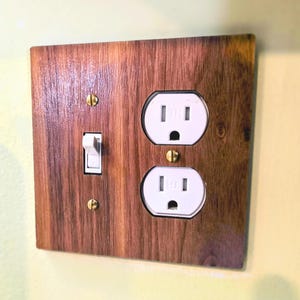 May include: A rectangular wall plate with a wood grain finish featuring one white toggle light switch and two white electrical sockets. The plate is secured with three brass-coloured screws, positioned vertically along the centre and left side.