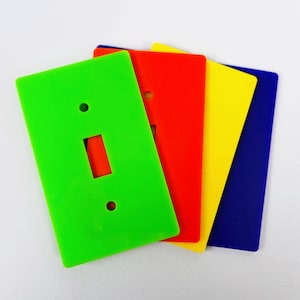May include: A stack of colorful light switch plates in green, red, yellow, and blue. Each plate has a rectangular opening for the switch and two small holes for mounting. The plates are made of a glossy plastic material.