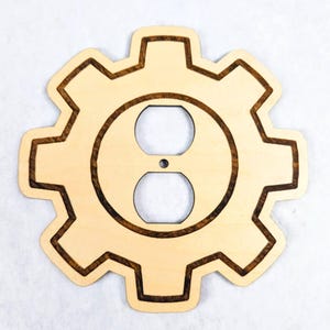 May include: A light-colored wooden wall plate cover shaped like a gear with eight teeth. The center features a standard duplex electrical outlet cutout with a small circular screw hole in the middle. The design includes a dark brown laser-etched border.
