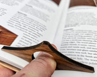 May include: A wooden book page holder in a dark brown finish. The holder is inserted into a book, with a finger hole for easy use. The book is open, displaying text.