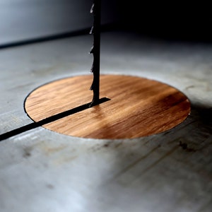 May include: Close-up of a band saw blade emerging from a wooden insert on a metal surface. The blade is black and serrated, and the wood is a warm brown color. The saw is likely used for woodworking.