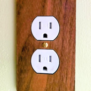 May include: A rectangular wall plate with a brown wood grain finish featuring two white electrical sockets stacked vertically. A single brass-coloured screw is positioned in the centre between the two sockets to secure the plate to the wall.