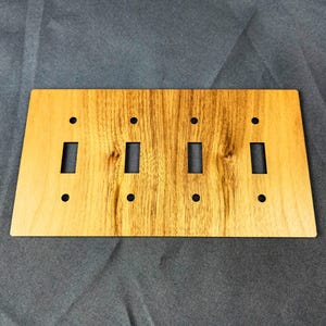 May include: A wooden light switch plate with four switch openings and four screw holes. The plate has a natural wood grain pattern with shades of brown and tan. The plate is rectangular with a slightly angled bottom edge.