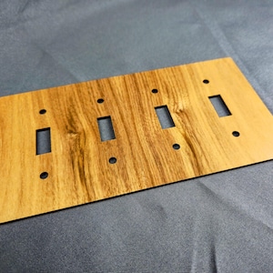 May include: A rectangular light switch wall plate with a wood grain pattern in shades of tan and brown. The plate features four vertical rectangular cutouts for toggle switches and eight small circular holes for mounting screws.