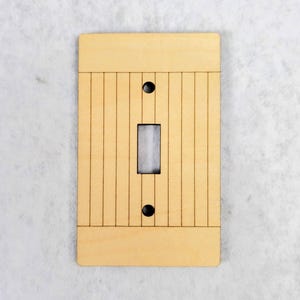 May include: A rectangular light-colored wooden light switch cover plate featuring a vertical slat pattern. The plate includes a central rectangular cutout for a toggle switch and two circular holes for mounting screws at the top and bottom.