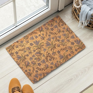 Puede incluir: Una alfombra rectangular con un estampado floral en tonos marrones, verdes y morados. La alfombra está colocada frente a una puerta, con una cesta y zapatos visibles en el fondo. El diseño presenta flores y hojas estilizadas.