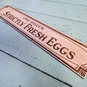 Op de afbeelding: Houten bord met de tekst "Strictly Fresh Eggs, One Dozen" in een decoratief lettertype. De tekst en randen zijn donkerbruin. Het rechthoekige bord heeft afgeronde uiteinden.