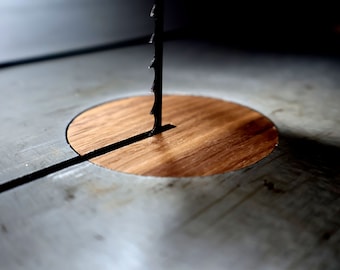 Puede incluir: Primer plano de una hoja de sierra de cinta que emerge de una pieza de madera en una superficie metálica. La hoja negra está dentada y la madera es de color marrón cálido. La imagen sugiere un taller de carpintería.