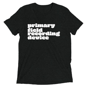 Peut inclure: T-shirt gris foncé avec l'inscription blanche "primary field recording device" superposée. Le t-shirt a des manches courtes et un col rond.