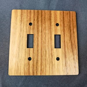 Op de afbeelding: Houten dubbele lichtschakelaarplaat met een natuurlijke houtnerf afwerking. De plaat heeft twee rechthoekige openingen voor de schakelaars en vier kleine ronde gaten voor montageschroeven. Het hout heeft een warme, lichtbruine kleur.