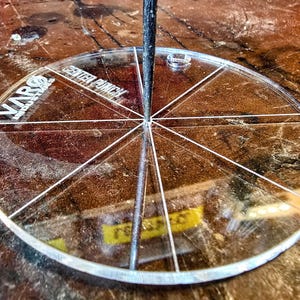 May include: A clear, circular VARO Industries center punch guide with white markings and text. The guide is divided into segments by white lines radiating from the center. A black rod is inserted through the center of the guide.