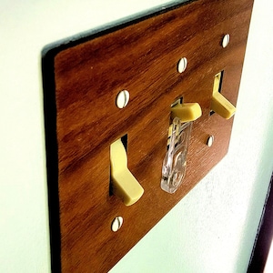 May include: A rectangular light switch wall plate with a brown wood grain finish. The plate features three toggle switches in a cream colour, with one switch equipped with a clear plastic safety guard. It is secured by four white screws.