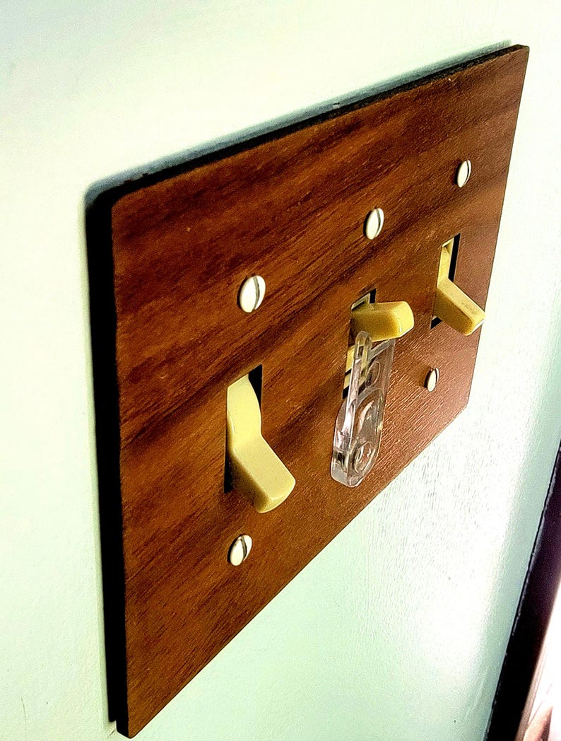 May include: A rectangular light switch wall plate with a brown wood grain finish. The plate features three toggle switches in a cream colour, with one switch equipped with a clear plastic safety guard. It is secured by four white screws.