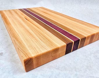 May include: A rectangular wooden chopping board with a light wood grain. It features a central stripe of purple and brown wood, flanked by lighter and darker wood tones. The board is approximately 40 cm long and 30 cm wide.
