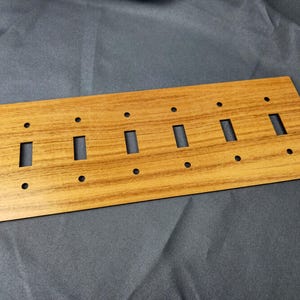 May include: A wooden light switch plate with a wood grain effect. It features six rectangular switch apertures and six small circular screw holes. The wood has a warm, natural tone, similar to teak.