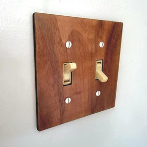 May include: A double light switch wall plate made of brown wood with a visible grain pattern. The rectangular plate features two cream-coloured toggle switches positioned vertically and is secured to the wall with four silver-coloured metal screws.