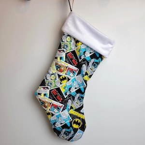 Puede incluir: Una media de Navidad negra con un puño blanco. La media está decorada con un patrón de cómic con Batman y el texto "BATMAN".