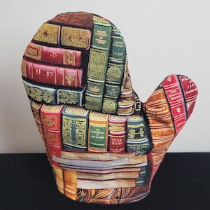 Puede incluir: Una manopla de horno marrón y roja con un diseño de estampado de libros. La manopla tiene un mango largo y una punta puntiaguda. Los libros de la manopla tienen letras doradas y están dispuestos en un patrón de estantería.