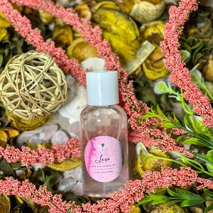 Puede incluir: Una botella de vidrio transparente con una tapa blanca y una etiqueta rosa y blanca que dice "Love Fire Girl Crystal Oil". La botella está rodeada de flores secas y otros elementos naturales.