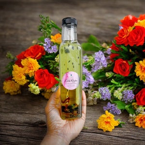 Może przedstawiać: Przezroczysta szklana butelka z zielonym płynem i etykietą z napisem "Herb Magic" Olejek do włosów wzbogacony ziołami. Butelka jest trzymana przez rękę przed bukietem kolorowych kwiatów.