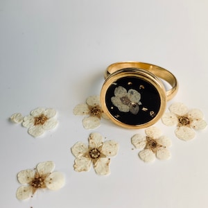 Peut inclure: Une bague en or avec un sertissage circulaire noir orné d'une fleur blanche pressée. La bague est entourée de petites fleurs blanches.