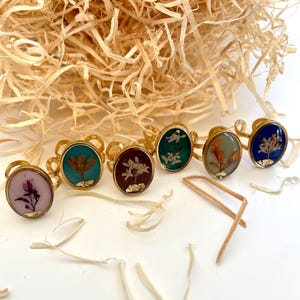 Op de afbeelding: Zes gouden ringen met verschillende gekleurde harsinlegsels met geperste bloemen. De ringen zijn in een halve cirkel op een wit oppervlak gerangschikt.