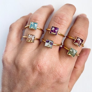 Op de afbeelding: Een set van zes gouden ringen met vierkante settings met geperste bloemen. De ringen zijn gestapeld op een hand, waardoor de verschillende bloemendesigns worden getoond.