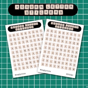 Op de afbeelding: Twee vellen houten letterstickers worden getoond op een groene rasterachtergrond. Elk vel heeft een zwarte rand en bevat individuele lettertegels in een lettertype dat op Scrabble lijkt. De bovenkant van elk vel leest "WOODEN LETTER STICKERS".