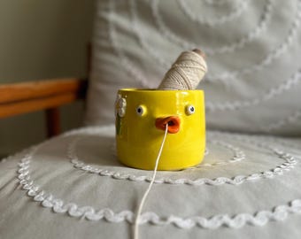 以下が含まれることがあります： 2つの小さな目とオレンジ色のくちばしを持つ、鳥の形をした鮮やかな黄色の陶器製ヤーンボウル。オフホワイトの毛糸玉がボウルの中に入っており、毛糸の端がくちばしの開口部から引き出されています。