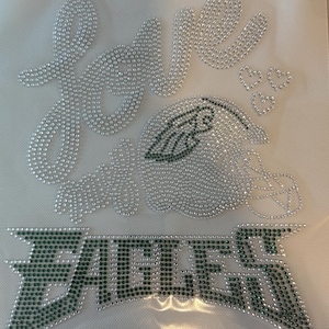 Puede incluir: Un diseño con la palabra "love" y la palabra "EAGLES" en escritura cursiva, un casco de fútbol y tres corazones. El diseño está hecho de pedrería plateada y verde sobre un fondo blanco.