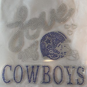 May include: A white fabric with the word "love" and the word "Cowboys" spelled out in silver and blue rhinestones. A blue rhinestone football helmet is also featured.