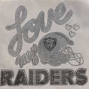Könnte beinhalten: Ein weißer Stoff mit einem Design aus silbernen Strasssteinen. Das Design umfasst das Wort "love" in Schreibschrift, einen Footballhelm, drei Herzen und das Wort "Raiders" in Blockbuchstaben. Das Design ist in Silber und Schwarz.