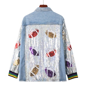 May include: Light blue denim jacket with a sequined back panel featuring colorful footballs. The jacket has a classic collar, long sleeves, and a ribbed cuff with green, purple, and yellow stripes. The jacket is hanging from a wooden hanger.