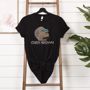 May include: A black short-sleeve t-shirt featuring a rhinestone graphic of a person wearing blue sunglasses. The design includes the text CHRIS BROWN in silver rhinestones below the portrait. The shirt has a crew neckline and a standard fit.