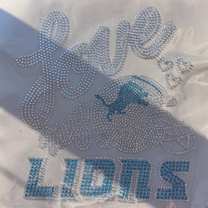 Pode incluir: Um design de transferência de strass numa superfície de tecido branco. A palavra love está escrita em cristais transparentes sobre um gráfico de capacete de futebol com um logótipo de leão azul. A palavra LIONS está em cristais azuis.
