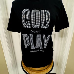 Puede incluir: Camiseta negra con un diseño de pedrería que dice "GOD DON'T PLAY ABOUT ME".