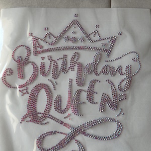 May include: A rhinestone design on a mesh backing. The design features the words "Birthday Queen" in a script font, topped with a crown. The rhinestones are primarily pink and iridescent, creating a sparkling effect. Ideal for DIY projects.