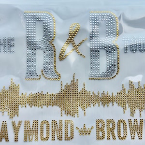 May include: A rhinestone iron-on transfer featuring the text THE R&B TOUR in silver and gold, a gold sound wave graphic, and the text RAYMOND BROWN with a small crown icon in gold. The design is set against a clear, flexible plastic backing.