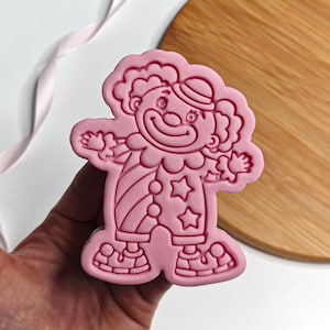 May include: Pink silicone mold of a cartoon clown with a red nose, a hat, and a striped outfit. The mold is designed for baking and decorating cookies or other treats.